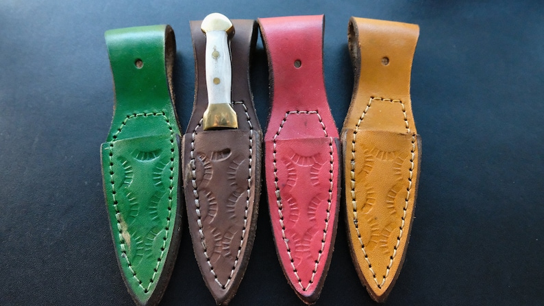 May include: Four small knives with sheaths in green, brown, red, and tan. Each knife has a white handle and a gold-coloured bolster. The sheaths are made of leather with decorative stitching and embossed patterns.