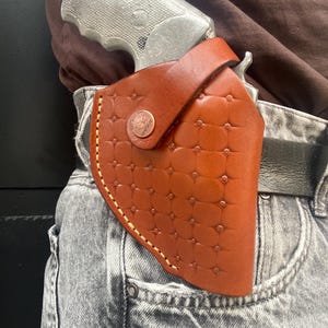 Puede incluir: Una funda de cuero color canela con un patrón en relieve estilo acolchado y un cierre de botón a presión color cobre. La funda está asegurada a un cinturón negro y sostiene un revólver metálico plateado, posicionado sobre jeans grises.