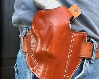 Könnte beinhalten: Eine braune Lederholster mit weißen Nähten ist an einem braunen Gürtel befestigt. Die Holster wird auf der rechten Seite getragen, und ein silbernes Objekt ist teilweise darin sichtbar. Die Person trägt eine blaue Jeans und ein blaues Langarmshirt.