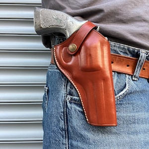 Puede incluir: Una funda de cuero marrón con una pistola plateada sujeta a un cinturón marrón. La funda se lleva en el lado derecho, sujeta a unos vaqueros azules. La funda tiene un cierre de botón a presión y costuras blancas.