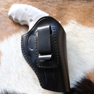 May include: A black leather holster with a metal clip, designed to hold a handgun. The holster is shown against a backdrop of brown and white fur.