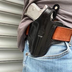 May include: A black leather holster with a silver handgun is attached to a brown belt. The holster is worn on the right side, visible against a person's blue jeans and grey shirt. The holster is designed to securely hold the firearm.