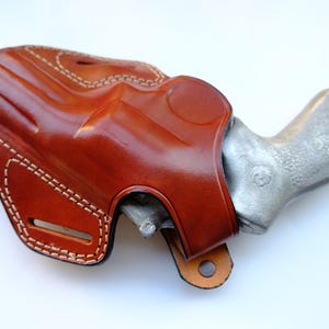 May include: A brown leather holster with white stitching, holding a silver handgun. The holster has a glossy finish and a loop at the bottom. The handgun's grip is visible.