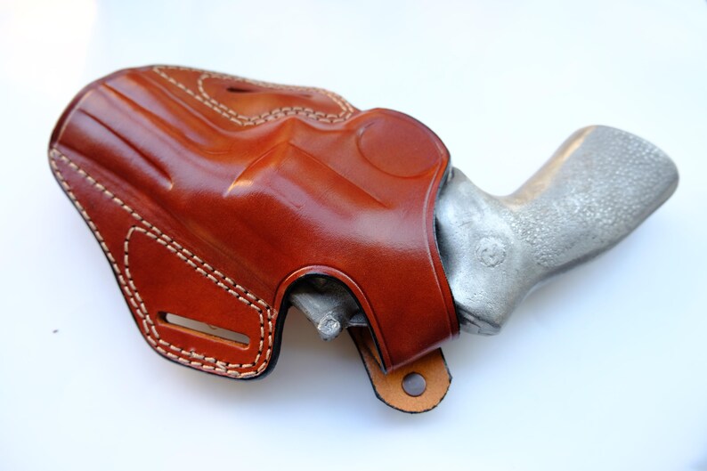 May include: A brown leather holster with white stitching, holding a silver handgun. The holster has a glossy finish and a loop at the bottom. The handgun's grip is visible.