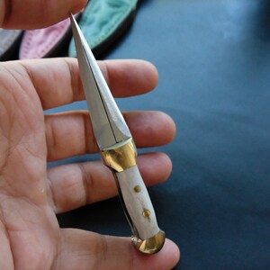 May include: A small, handcrafted knife with a silver blade and a white handle. The handle has brass fittings. The knife is held in a hand, showing its size and detail. The blade is sharp and pointed.