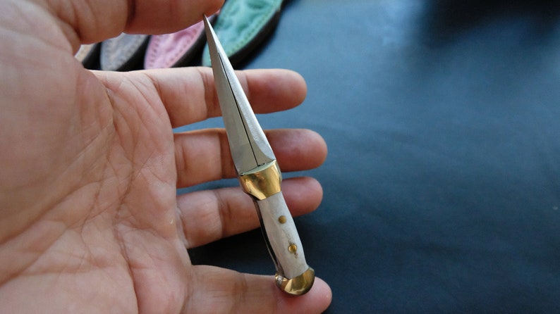May include: A small, handcrafted knife with a silver blade and a white handle. The handle has brass fittings. The knife is held in a hand, showing its size and detail. The blade is sharp and pointed.