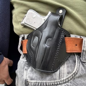 May include: Black leather holster with a silver handgun. The holster is worn on a brown belt, attached to grey jeans. The holster has a molded design and is stitched along the edges.