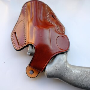May include: A brown leather holster with a stitched design, holding a silver handgun. The holster has a glossy finish and a loop for a belt. The handgun's grip is textured.