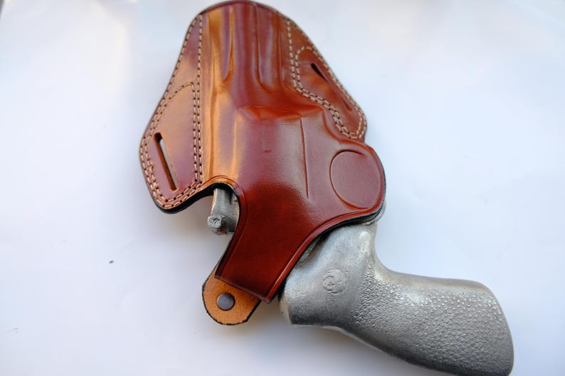 May include: A brown leather holster with a stitched design, holding a silver handgun. The holster has a glossy finish and a loop for a belt. The handgun's grip is textured.