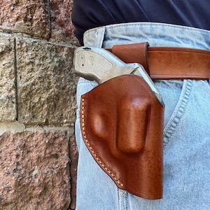 Peut inclure: Un étui en cuir marron contenant un revolver de couleur argentée, fixé à une ceinture en cuir marron portée sur un jean en denim bleu clair. L'étui présente des coutures visibles sur le bord et est placé sur la hanche contre un mur de briques.