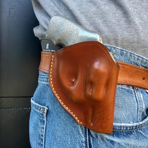 May include: A brown leather holster with a light-colored stitched border, worn on a belt with a silver handgun inside. The holster is attached to a brown leather belt, and the person is wearing blue jeans and a gray shirt.