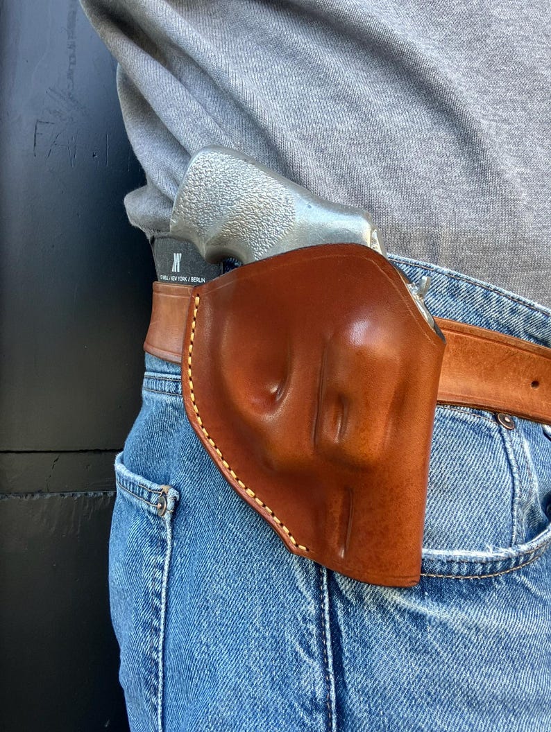 May include: A brown leather holster with a light-colored stitched border, worn on a belt with a silver handgun inside. The holster is attached to a brown leather belt, and the person is wearing blue jeans and a gray shirt.