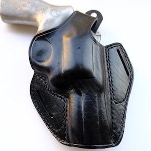 May include: A black leather holster with a silver-colored handgun. The holster is molded to the shape of the gun and features detailed stitching. The leather has a glossy finish, and the design includes a belt loop.