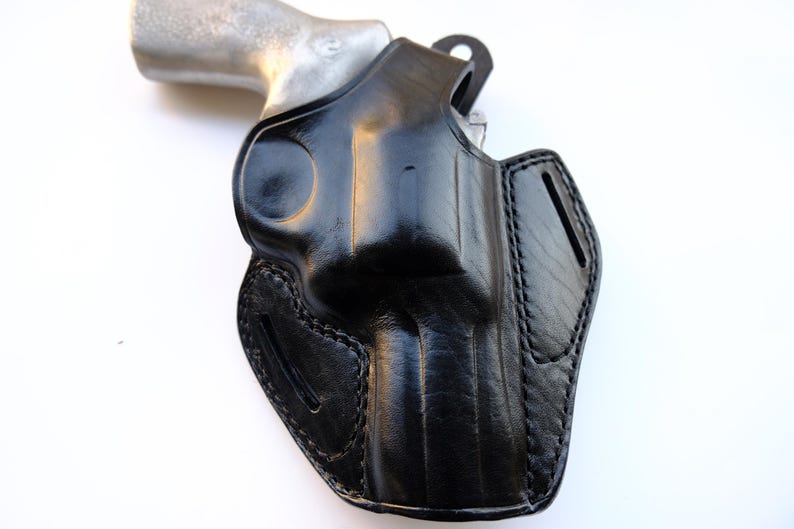 May include: A black leather holster with a silver-colored handgun. The holster is molded to the shape of the gun and features detailed stitching. The leather has a glossy finish, and the design includes a belt loop.