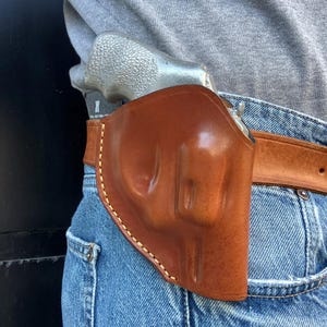 May include: A brown leather holster with a light brown belt, worn on blue jeans. The holster is designed to hold a silver revolver. The holster is stitched with a light-colored thread.