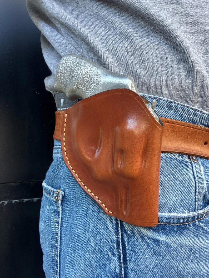 May include: A brown leather holster with a light brown belt, worn on blue jeans. The holster is designed to hold a silver revolver. The holster is stitched with a light-colored thread.