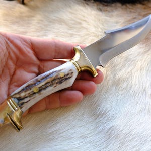 May include: A fixed-blade knife featuring a polished silver-toned steel blade, a brass bolster, and a textured handle made of natural antler material. The handle is secured with three brass pins and features a brass pommel at the base.