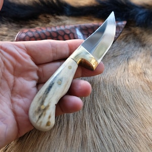 Peut inclure: Un couteau à lame fixe doté d'une lame en acier poli, d'une mitre en laiton et d'un manche texturé de couleur crème fixé par trois goupilles en laiton. Un étui en cuir marron à motif nid d'abeille est placé derrière le manche du couteau.