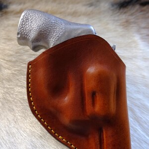 May include: A brown leather holster with yellow stitching, holding a silver handgun. The holster is made of smooth, polished leather. The handgun's grip is visible, with a textured surface.