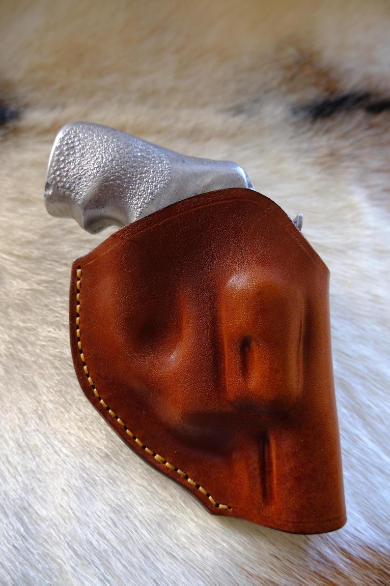 May include: A brown leather holster with yellow stitching, holding a silver handgun. The holster is made of smooth, polished leather. The handgun's grip is visible, with a textured surface.