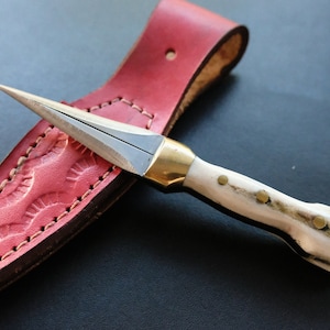 May include: A handmade dagger with a sharp, silver blade and a bone handle, accented with brass fittings. The dagger rests in a red leather sheath with decorative stitching. The overall length of the dagger is approximately 7 inches.