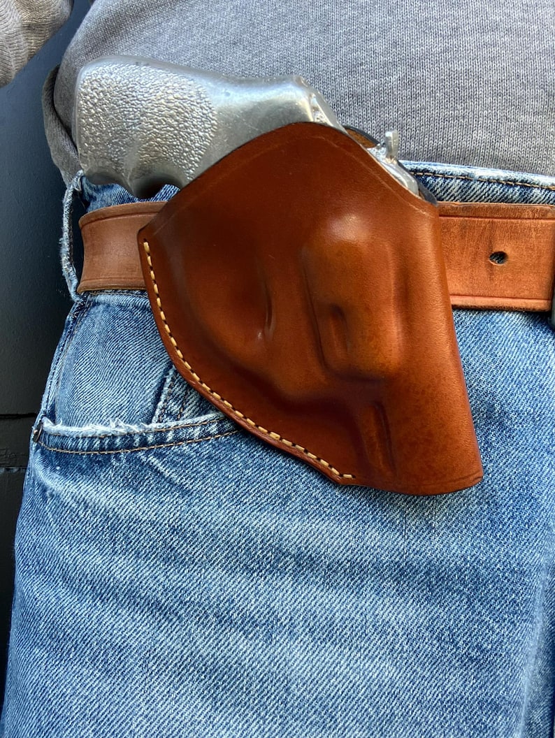 May include: A brown leather holster with white stitching, holding a silver handgun. The holster is attached to a brown leather belt, worn over blue denim jeans. The image is a close-up shot.