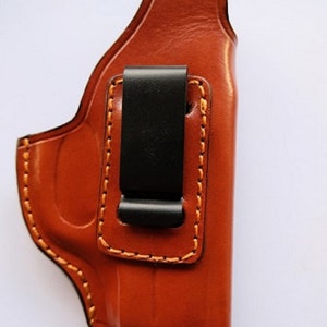 Puede incluir: Una funda de cuero marrón con un clip negro. La funda tiene costuras de color naranja y está diseñada para sostener una pistola.