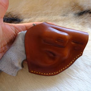 May include: A brown leather holster with yellow stitching, holding a silver and gray handgun. The holster is molded to the shape of the gun. The image is set against a background of fur.