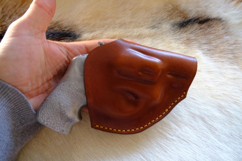 May include: A brown leather holster with yellow stitching, holding a silver and gray handgun. The holster is molded to the shape of the gun. The image is set against a background of fur.