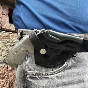 May include: A black leather holster containing a silver-coloured revolver, secured with a single silver snap button. The holster features decorative stitching along the bottom edge and is attached to a black belt worn over grey denim jeans.