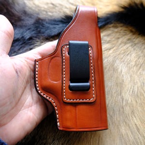 Puede incluir: Una funda de cuero marrón con costuras blancas y un clip negro. La funda se sostiene en una mano, mostrando su diseño y artesanía. El cuero tiene un tono rico y cálido.