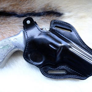 May include: A black leather holster with a silver revolver. The holster is designed to fit the gun securely, with visible stitching and belt loops. The revolver's grip has a textured pattern. The background is a mix of brown and white.