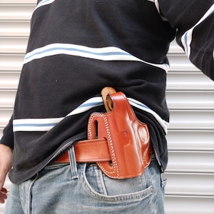 Op de afbeelding: Een bruine leren holster met een gestikt ontwerp, gedragen op de taille van een persoon met een riem. De holster is ontworpen om een ​​vuurwapen te dragen.