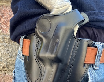 Könnte beinhalten: Eine schwarze Lederholster mit einem silbernen Revolver. Die Holster ist an einem braunen Gürtel befestigt, der mit einer Jeans getragen wird. Die Holster hat detaillierte Nähte.