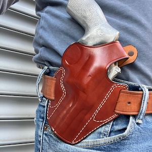 May include: A brown leather holster with white stitching, worn on a brown leather belt. A silver handgun is partially visible inside the holster. The person is wearing blue jeans and a blue long-sleeve shirt.