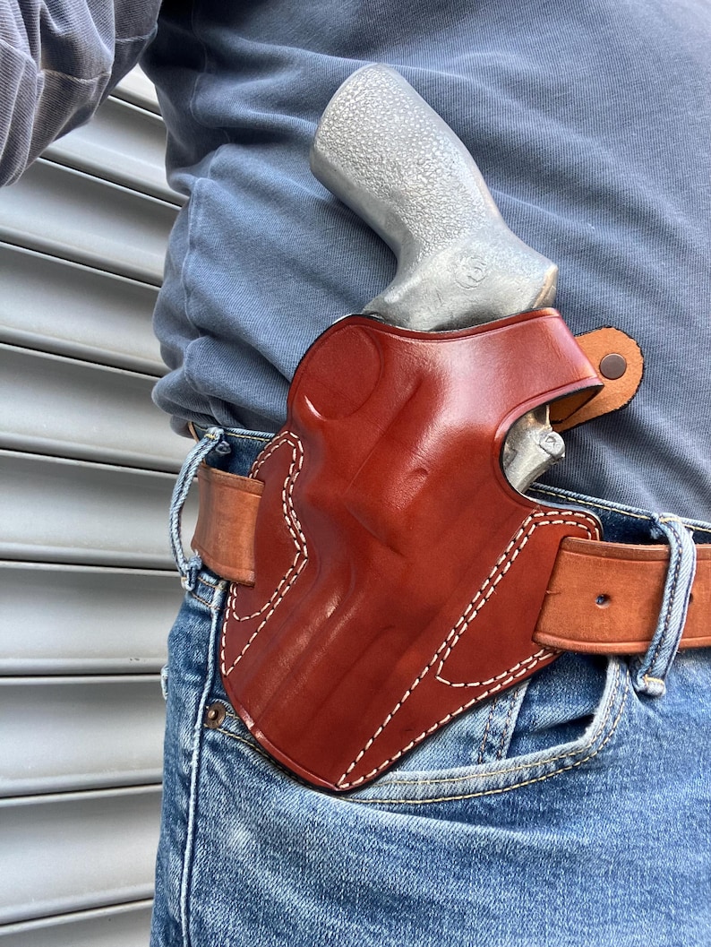 May include: A brown leather holster with white stitching, worn on a brown leather belt. A silver handgun is partially visible inside the holster. The person is wearing blue jeans and a blue long-sleeve shirt.