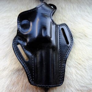 Puede incluir: Una funda de cuero negro con un acabado brillante. La funda tiene una forma moldeada con bordes cosidos y ranuras. El fondo es una superficie clara y texturizada.