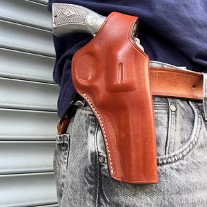 Könnte beinhalten: Ein braunes Lederholster mit weißen Nähten, zur Aufnahme eines Revolvers. Das Holster ist an einem braunen Ledergürtel befestigt, der mit grauen Jeans getragen wird. Der Revolver ist teilweise sichtbar.