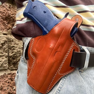 Puede incluir: Una funda de cuero marrón con costuras blancas, que sostiene una pistola azul. La funda está sujeta a un cinturón negro que se lleva con vaqueros azules. La pistola tiene un agarre texturizado.