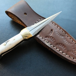 May include: A small, handcrafted knife with a sharp, pointed blade and a white handle with black accents. The knife features gold-coloured metal accents and rests on a brown leather sheath with decorative stitching and embossed design.