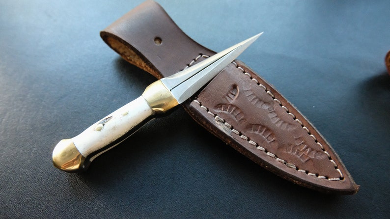 May include: A small, handcrafted knife with a sharp, pointed blade and a white handle with black accents. The knife features gold-coloured metal accents and rests on a brown leather sheath with decorative stitching and embossed design.