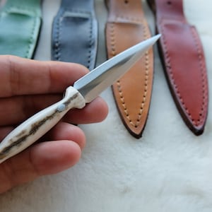 以下が含まれることがあります： 白い柄と銀色の刃を持つ小さなナイフ。ナイフは手で持たれており、背景には革製の鞘がいくつかある。