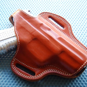 Puede incluir: Una funda de cuero marrón con costuras blancas para una pistola. La funda tiene dos trabillas para el cinturón y una protección para el gatillo.