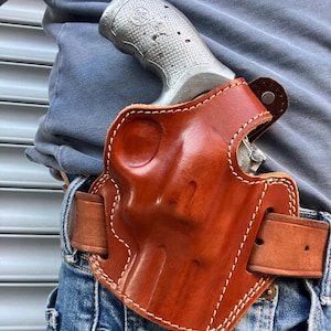 Puede incluir: Una funda de cuero marrón con costuras blancas, que sostiene un revólver plateado. La funda está sujeta a un cinturón de cuero marrón que se lleva con vaqueros azules. Se ve la empuñadura del revólver.