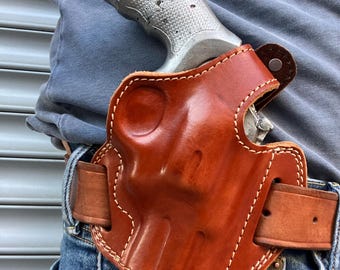 May include: A brown leather holster with white stitching, holding a silver revolver. The holster is attached to a brown leather belt worn with blue jeans. The revolver's grip is visible.
