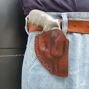 Op de afbeelding: Een bruine lederen holster met decoratieve geschulpte randen en rode stiksels, gedragen aan een bruine lederen riem over een lichtblauwe spijkerbroek. De holster bevat een zilverkleurige revolver met een textuurhandgreep en een logo.