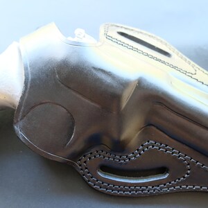 Può includere: Un fodero in pelle nera per una pistola. Il fodero ha un passante per la cintura e un fermo per il pollice.