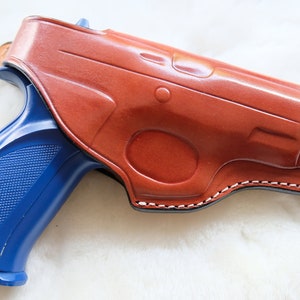 Puede incluir: Una funda de cuero marrón con una pistola azul en su interior. La funda tiene un borde cosido y una solapa que cubre la protección del gatillo.