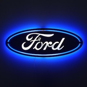May include: A neon sign displaying the Ford logo. The sign has an oval shape with a dark blue background and a bright blue border. The word "Ford" is written in white, stylized script.