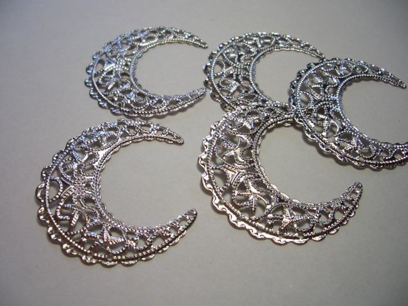 May include: Four silver-toned metal crescent moon charms featuring an intricate filigree cutout pattern. Each piece has a scalloped edge and a reflective metallic finish, arranged in a cluster to display the detailed openwork design.